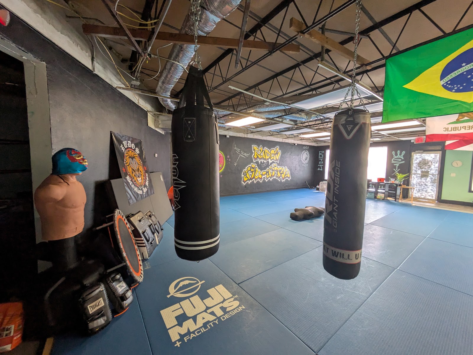 Wide view of the Rudos BJJ facility showing royal blue Fuji mats, heavy bags, Lucha Libre masked striking dummy, and graffiti mural