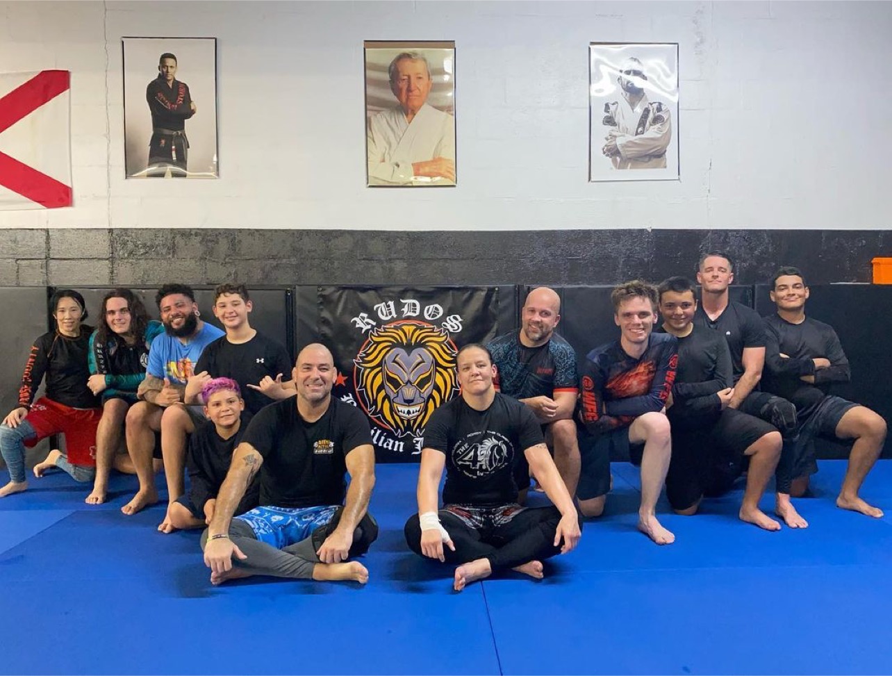 Full No-Gi room seated in front of the Rudos Lion crest with Prof. Shayna Baszler and Prof. David.